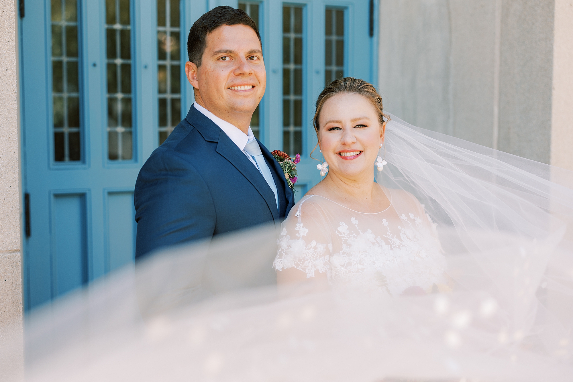 newlyweds stand together in front of blue windows at St. Ann’s Roman Catholic Church
