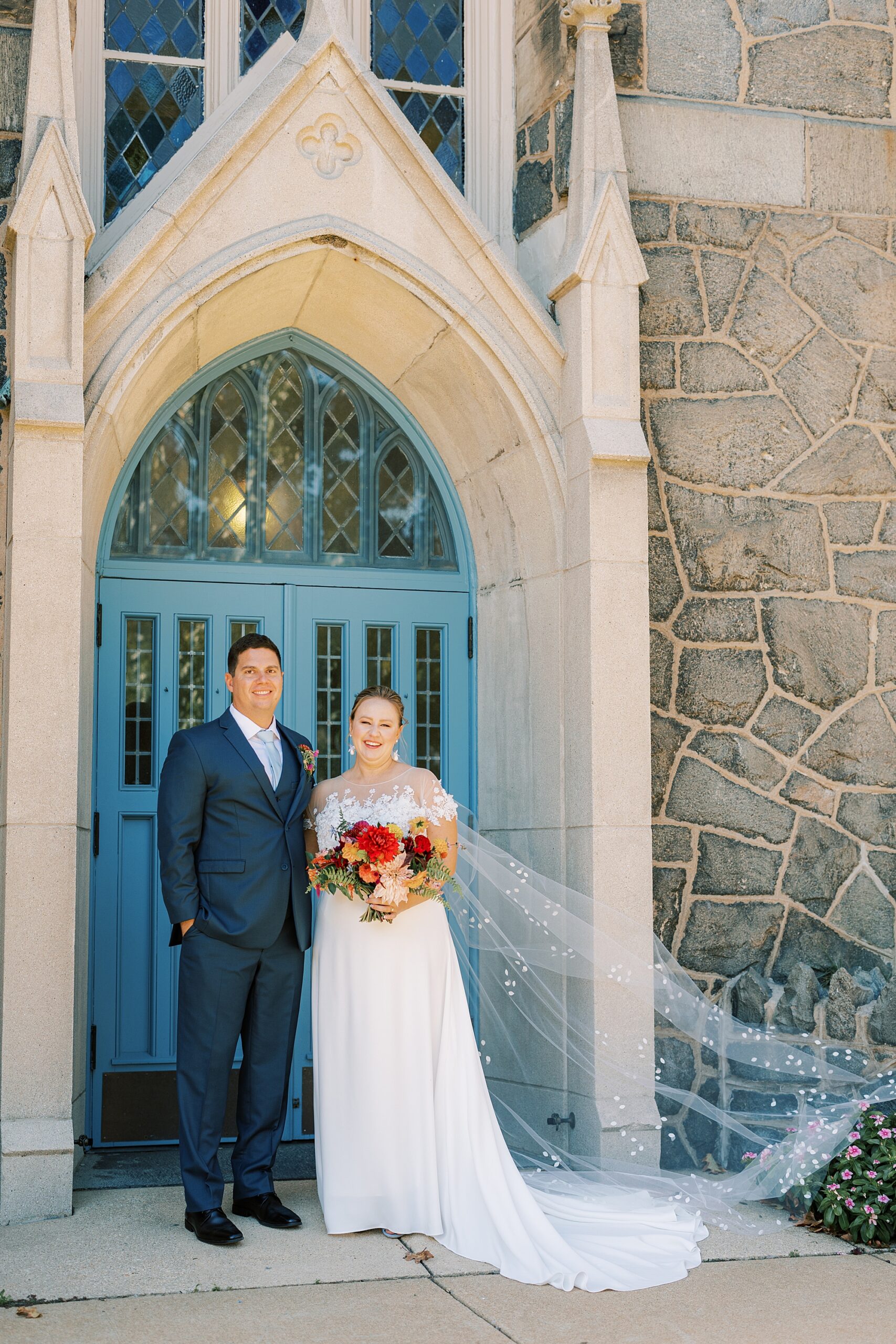 bride and groom stand by blue doors under stone arbor at St. Ann’s Roman Catholic Church
