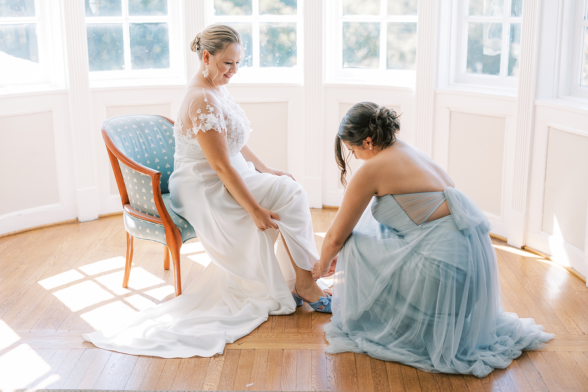 bridesmaid in blue dress helps bride with blue shoes in chair by window 