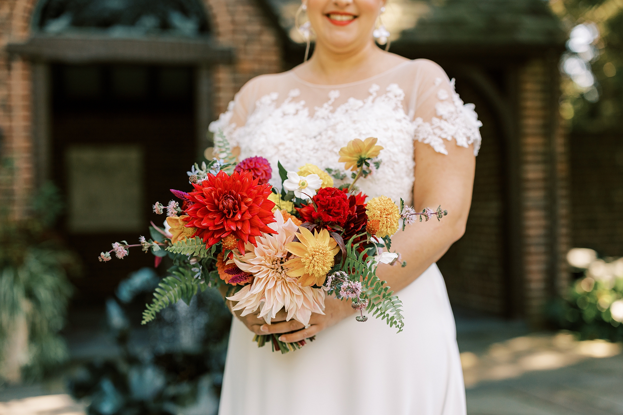 bride in wedding gown with lace cover holds bouquet of red and yellow flowers 