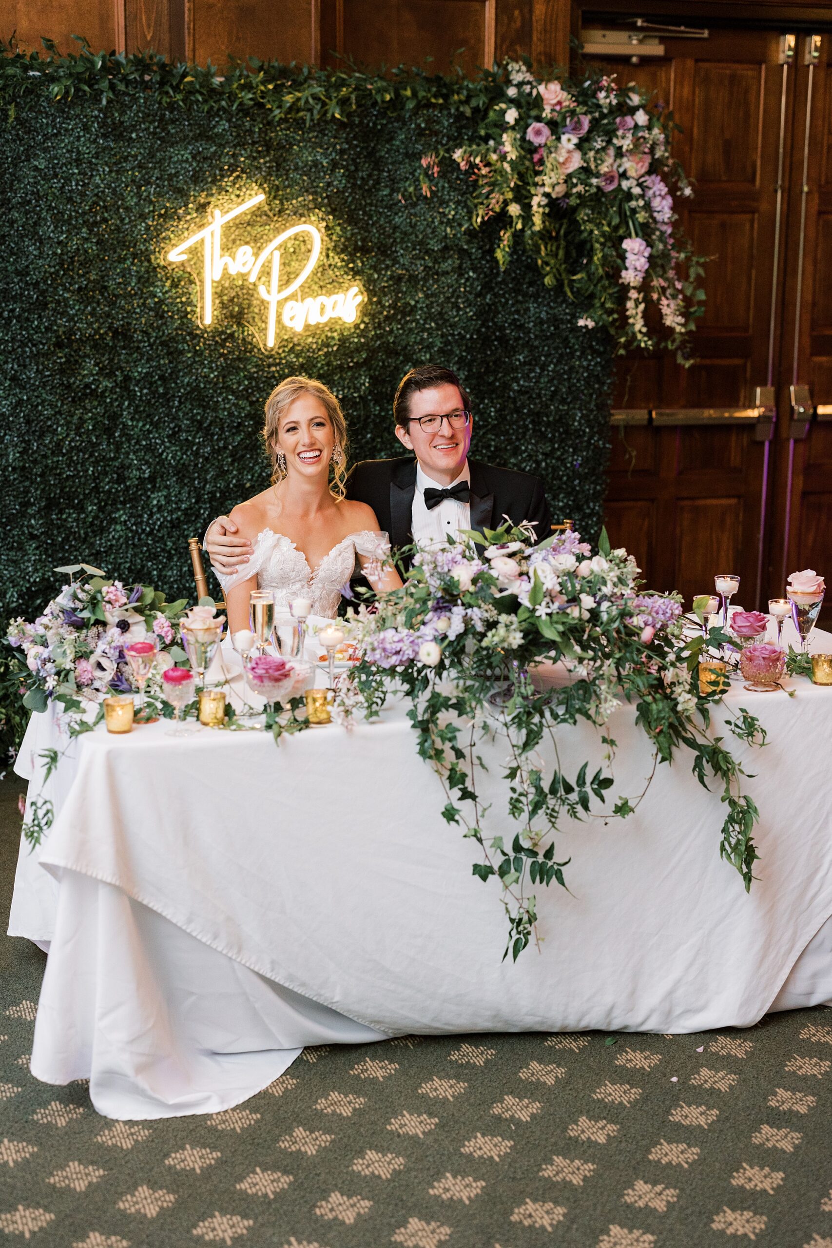 bride and groom sit at sweetheart table with greenery wall behind them