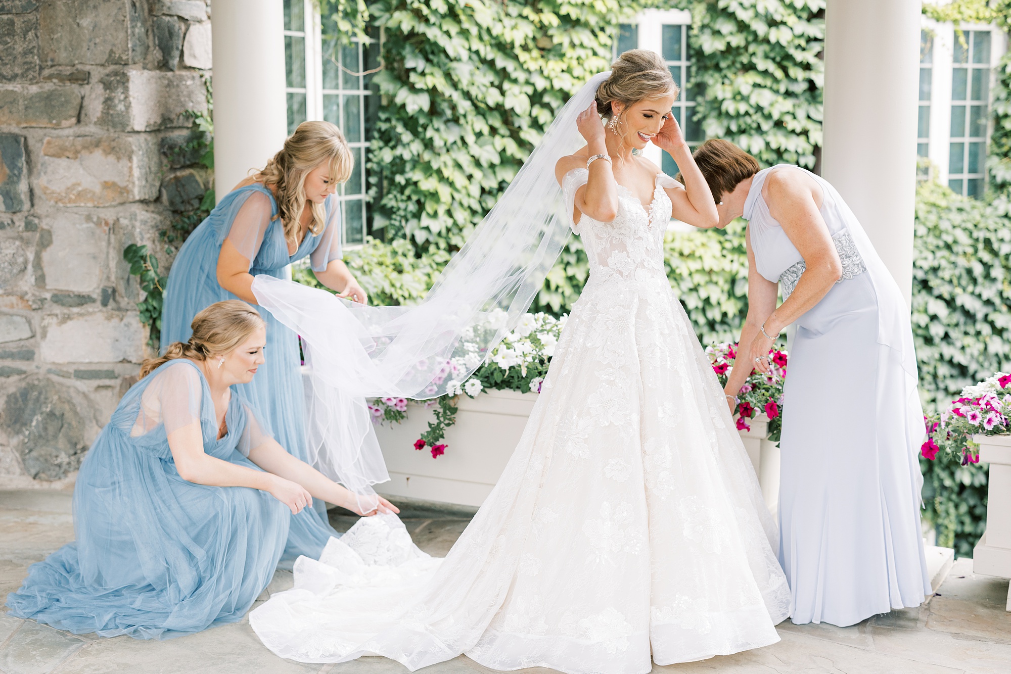bridesmaids in blue gowns help bride with dress and veil 
