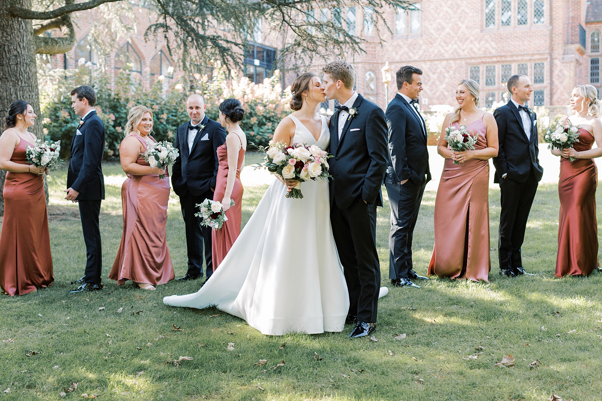bride and groom kiss with wedding party around them in burnt orange gowns and black suits 