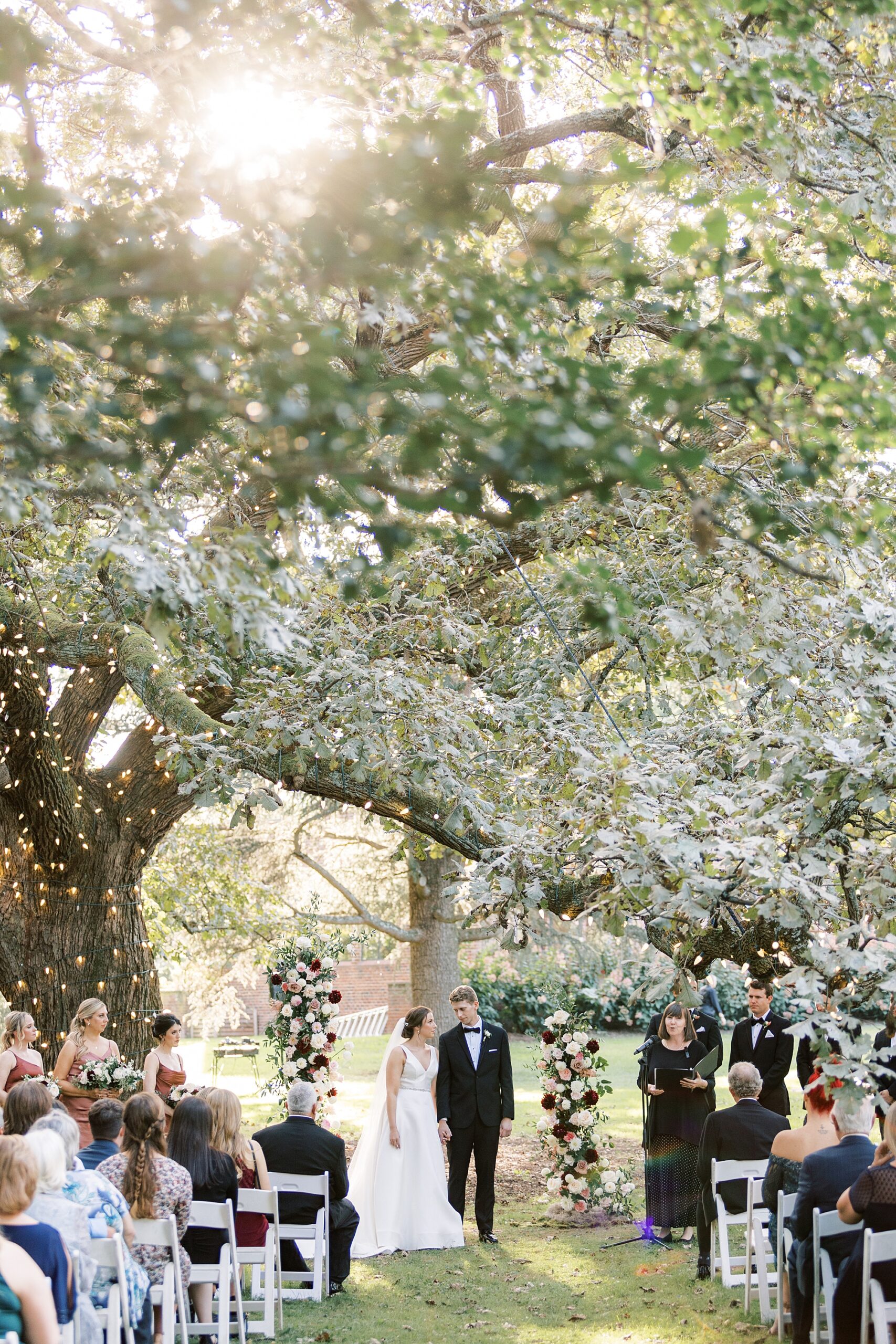 newlyweds hold hands under oak tree on lawn at Aldie Mansion during wedding ceremony 