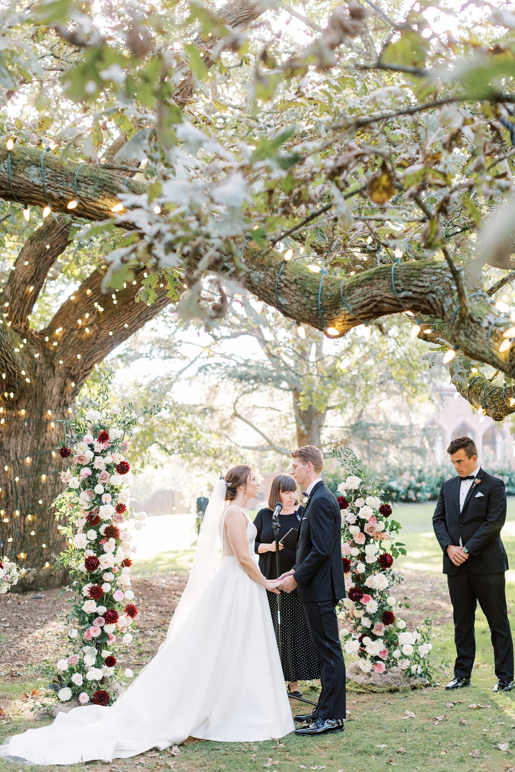 bride and groom hold hands by tall floral installations under tree at Aldie Mansion