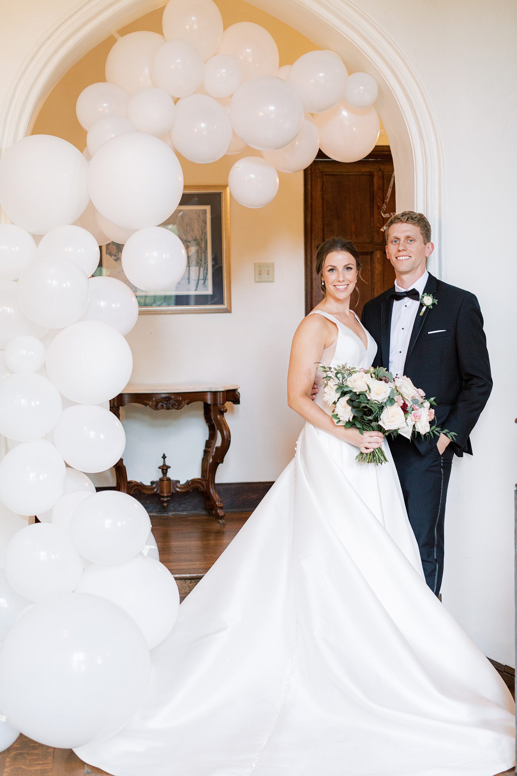 bride and groom lean against wall inside Aldie Mansion with ivory balloon arbor 
