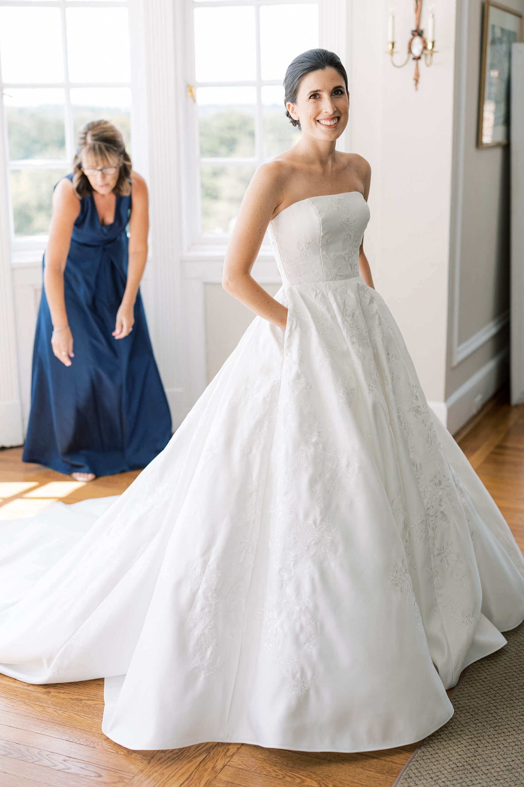 bridesmaid in navy gown helps bride into strapless wedding dress