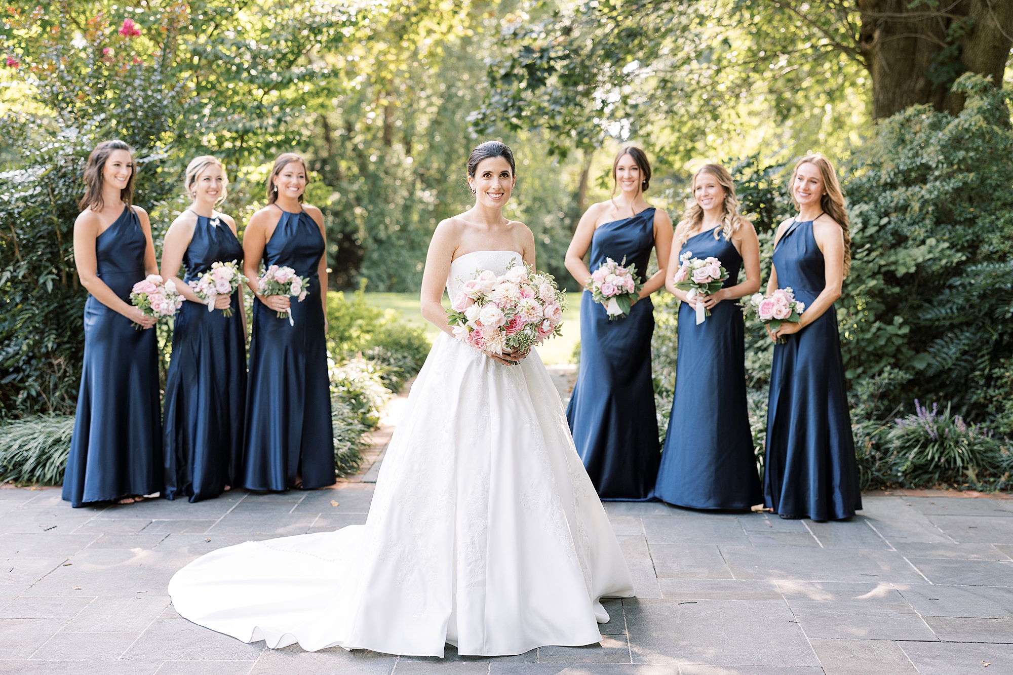 bride stands with bridesmaids in navy gowns with pink and white bouquets 