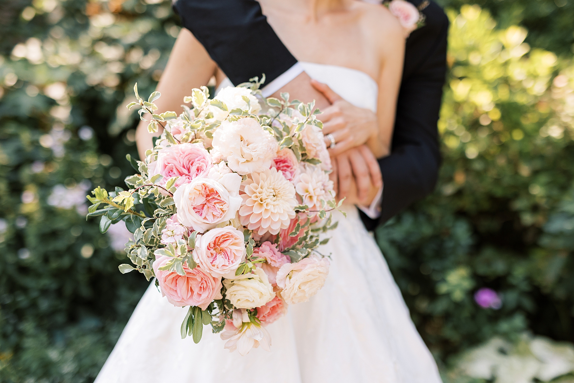groom hugs bride from behind while she holds bouquet of pink peonies 